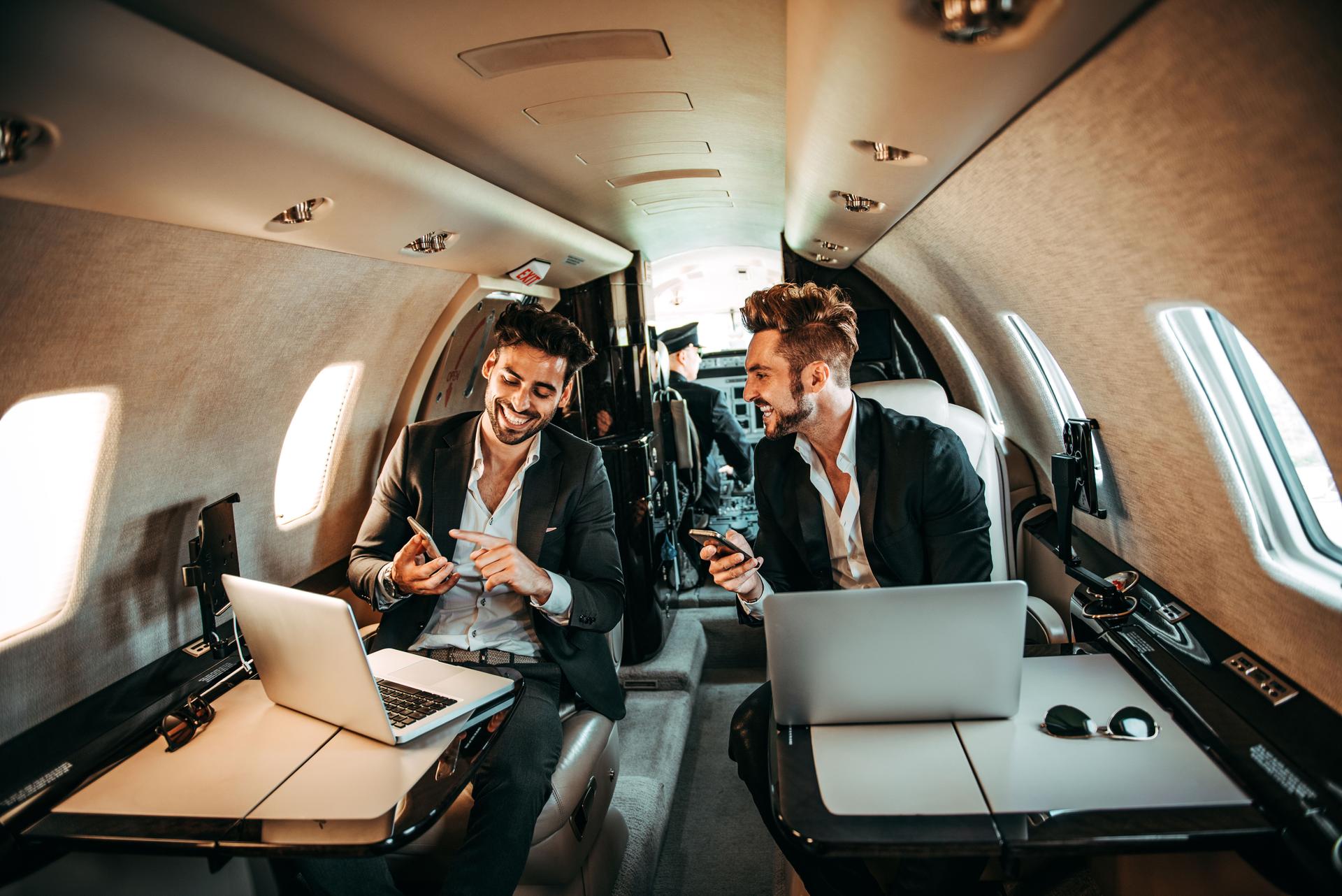 Two successful corporate managers using mobile phones while working on laptops together are flying on a private jet